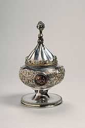 Silver gilt vessel with conical lid. It was intended for use as a ciborium. The vessel is decorated with foliate scrolls and medallions. The knob on the vessel's lid is in the shape of a bunch of grapes.