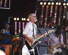 Gillies at left behind his drum kit. Johns is singing wide-mouthed into a microphone and playing his guitar, he wears a garter with a ribbon over his pants on his right thigh. Joannou is playing his bass guitar. In the background are three female singers with one partly obscured at right.