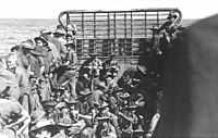 About forty troops wearing slouch hats and carrying rifles packed into a landing craft travelling across open water, viewed from the stern towards the bow ramp.