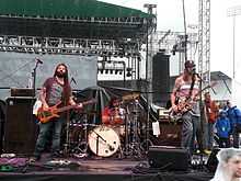 One male guitarist, one male bassist and one male drummer performing on an outdoor stage