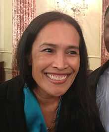 A photograph of a smiling woman with long, black hair looking to the right and wearing a necklace, a blue shirt, and a black suit jacket