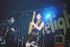 A female bass guitarist and male guitarist perform on stage, with stage lights and a backdrop that reads "The Limelight" in yellow on a black wall.