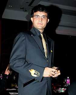 A clean-shaven man wearing glasses stares directly at the camera. He is wearing a black suit and a gold tie. The man's right arm is facing the camera. The arm has a small picture of a helmet engulfed with fire.