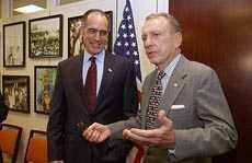 Two men wearing suits, in discussion with each other, standing in front of the United States flag and wall covered in framed photographs