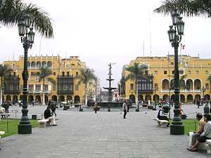 Large square with a fountain and four storied yellow buildings with large glassed balconies.