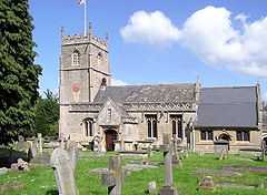 Yellow stone building, with porch with triangular roof in front. Short square tower with battelements toped by flag and falg pole. Gray gravestones in the foreground
