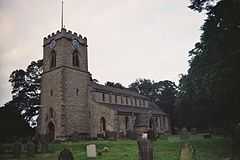 A small church with a square Ashlar tower, in the Early English style. The tower is to the left, the nave to the right, and we are looking somewhat obliquely at it.  The church is ringed by medium size trees, and a large Yew dominates the right of the picture, in the middle distance.  A handful of mismatched gravestones dot the grassy nearground of the picture.