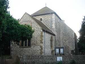 A flint rubble church with large stone quoins, behind a low flint wall. A three-light lancet window in the nearest wall is partly obscured by a tree in full leaf. A squat tower with a squared-off pyramidal roof stands in the middle. To its right is a blank-walled protrusion with a blocked entrance.