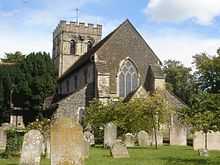 Rear view of a stone church with a castellated tower at the far end. The nearest side has very dark stone, two heavy buttresses and a three-light lancet window with trefoils. Trees surround the church on all sides, and there are several gravestones in front.