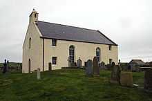 A large cream-coloured kirk, with two tall windows in the centre, a small belfry and a slate roof. The sea is visible in the background.
