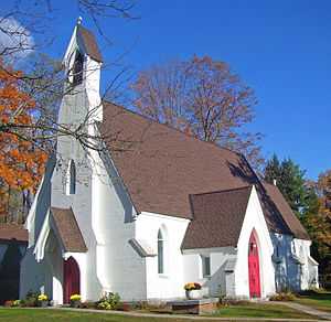 A white church with red pointed doors and a narrow belfry in front with a small cross on top. There is a low brick and stone structure in the foreground.