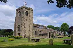 A stone church with a west tower, and a body of complex design including a porch and large rectangular paned windows in the clerestory.