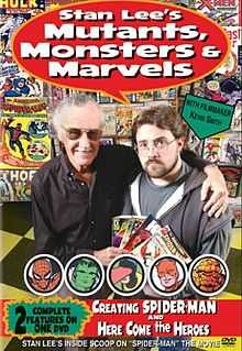 A photograph of two men, both of whom are wearing glasses, looking at the viewer, and standing on a yellow-and-black-checkered floor next to a wall of comic books