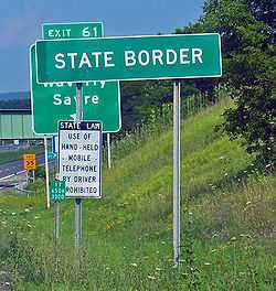 A white on green sign saying "State border". Behind it in the distance is a standard U.S. freeway exit sign.