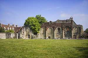 Landscape photo dominated by a field in front, surmounted by blue sky with white clouds. A ruined wall of a building crosses the center; in front a few stones can be seen piled atop each other.