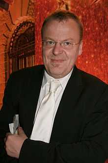 A photo of a bespectacled man wearing a black coat over a white collared shirt and white tie. He smiles.