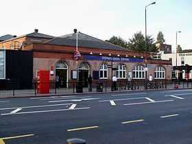 A brick building under a slate roof with a pale front; two arched doorways on the left and four arched windows to the right, above which a rectangular, dark blue sign reading "STEPNEY GREEN STATION" in white letters
