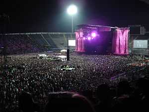 A stadium filled with spectators on the podiums and on the ground. In the middle is a stage with two giant pink 'M' symbols flanking it. A large flood light is visible behind it.