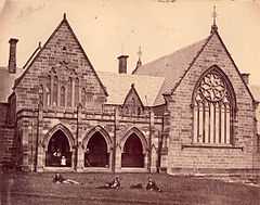 1870s photograph of church-like stone building, with students lying on the grass in front