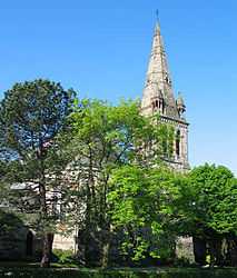 A stone-built church with a tower incorporating a spire, pictured against blue sky and largely obscured by a number of trees in full leaf.