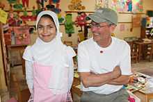 A photograph of a girl and a man sitting side-by-side, both smiling, the man looking at the girl and wearing a green baseball cap and a T-shirt