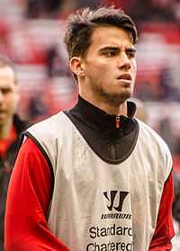 Close-up of a young man with brown hair, wearing a Liverpool-branded white training vest over a red tracksuit top