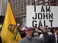 In a large outdoor crowd, a man holds up a poster with the words "I am John Galt" in all capital letters