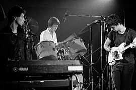 Three men are situated on a stage, each with his own respective instrument: a keyboard, a drum set and a guitar.