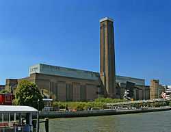 A large oblong brick building with square chimney stack in centre of front face. It stands on the far side of a river, with a curving white foot bridge on the left.