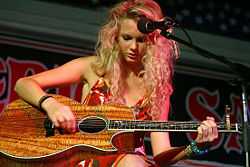 Taylor Swift sits and leans over her oak guitar while picking a string