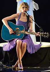 Taylor Swift, wearing a purple dress, plays a blue acoustic guitar while sitting on a stool