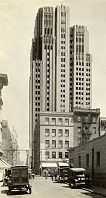 A monochrome photograph of a street in San Francisco showing automobiles from the 1920s, and a skyscraper standing tall behind smaller buildings.