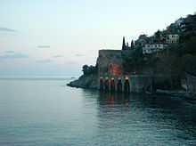 A hill populated with houses slants down into a blue-green sea below a stone dock with five arches. A stone wall extends along the sea from the dock to the lower right.