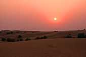 View of dunes at sunset. The setting sun hangs low and dim in a sky graded from fiery orange at top to maroon near the horizon. Terrestrial features are difficult to discern in the crepuscular illumination.