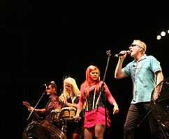 A group of musicians perform on a lighted stage. From left to right: a short-haired man plays bass guitar, a blond-haired woman plays the bongos, an red-haired woman holds a microphone, and a man wearing sunglasses and a blue shirt sings into a microphone.