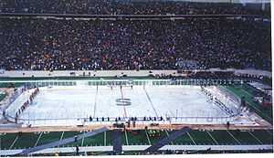 Two teams of many hockey players stand at either end of a rink built on top of a football field as thousands of people look on from the stands.