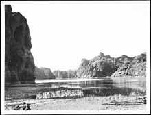 A steep rocky embankment looms in shadow at left. A tide mark is visible on the nearly vertical wall near the water line. The near bank is dried cracked mud. A few small pieces of driftwood sit on the bank. A rope lays on the bank at right. Across the placid river and in the distance rocky hills rise.