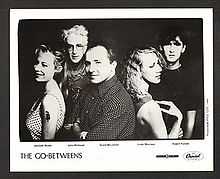 Five adults framed in upper body. In the front row, the left female is shown in left profile, slightly turned to her left and smiling, she wears a white dress with black polka dots and has a serpent tattoo on her upper left arm. Behind her, a slightly balding male is more turned towards the front and has his arms folded across his chest, his shirt is dark with white polka dots. Back to back to him is the second female in right profile with her right arm touching her shoulder. In the back row, the left male has white hair and is facing forward, he is wearing glasses and has an obscured design on his shirt. The right male has dark hair, he is staring forward and wears a black tee shirt.