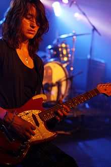 A woman performing live on-stage. She is holding a red guitar. A drum kit is seen in the background.