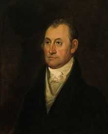 A man with dark, receding hair wearing a white shirt and black jacket