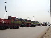 line of heavy trucks going north, background a container yard