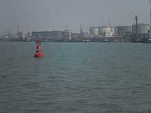 A red port-side buoy with an oil wharf and tankers in the background