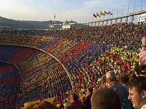 A crowd of spectators in an open-air stadium, a mosiac in red, blue and gold is visible