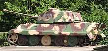 The side of a large tank, freshly painted in pale yellow, green and rust-brown camouflage, sitting in sunlight on a concrete plinth.