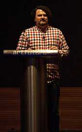 A middle aged Caucasian man with dark hair speaks from a lectern.