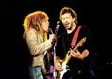 Turer and Clapton, on stage, sharing a microphone stand, singing.