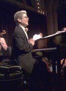 A smiling man with glasses sits at a piano on stage
