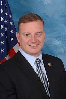 A middle-aged man wearing a dark blue suit, a light blue shirt, and a blue striped tie. In the background hangs an American flag.