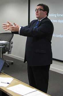 Large man in suit, glasses and short dark hair giving a presentation in front of overhead projection screen