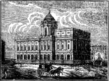 A neoclassical building seen from an angle.  It has two storeys and a dome on a high drum.  A horse and cart pass in front of it and to the right of the hall is a covered wagon